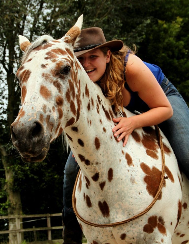 Vrouw in een leren cowboyhoed zit op een driekleurig gevlekt wit paard in de natuur met nekrope en omhelst het paard om de hals