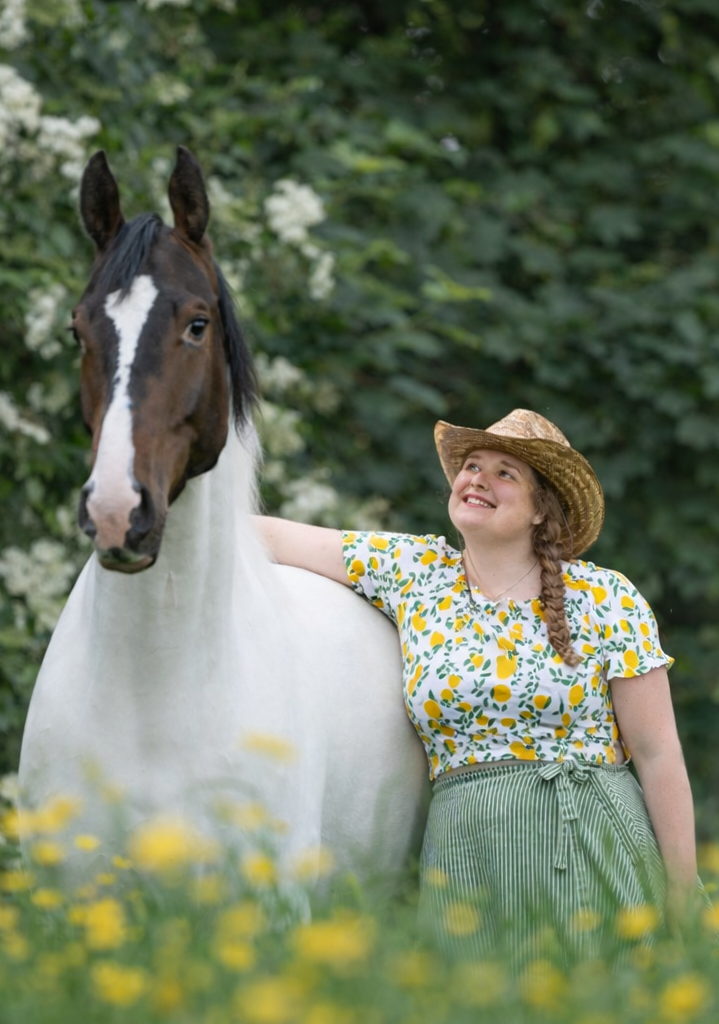 Michelle van MAGG Coaching kijkt vol trots naar Juno, haar paard, een bruin bonte KWPN ruin Vrouw in een witte blouse citroenen en rieten cowgirlhoed, staat naast paard in groen gras met gele bloemen. Heeft haar arm over zijn billen