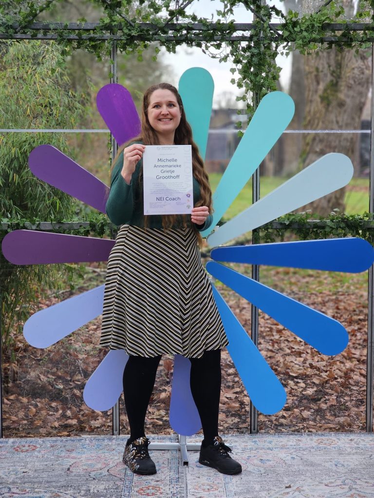Vrouw in groen vest en gestreepte rok houdt een diploma vast, achtergrond met kleurt logo van de opleiding Jolanda Vleugel.